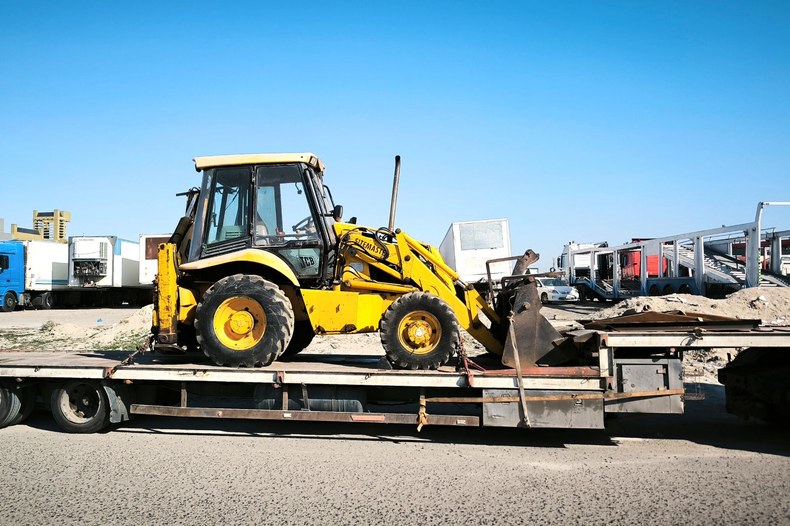 A yellow construction machine chained down on a flatbed trailer in an industrial yard