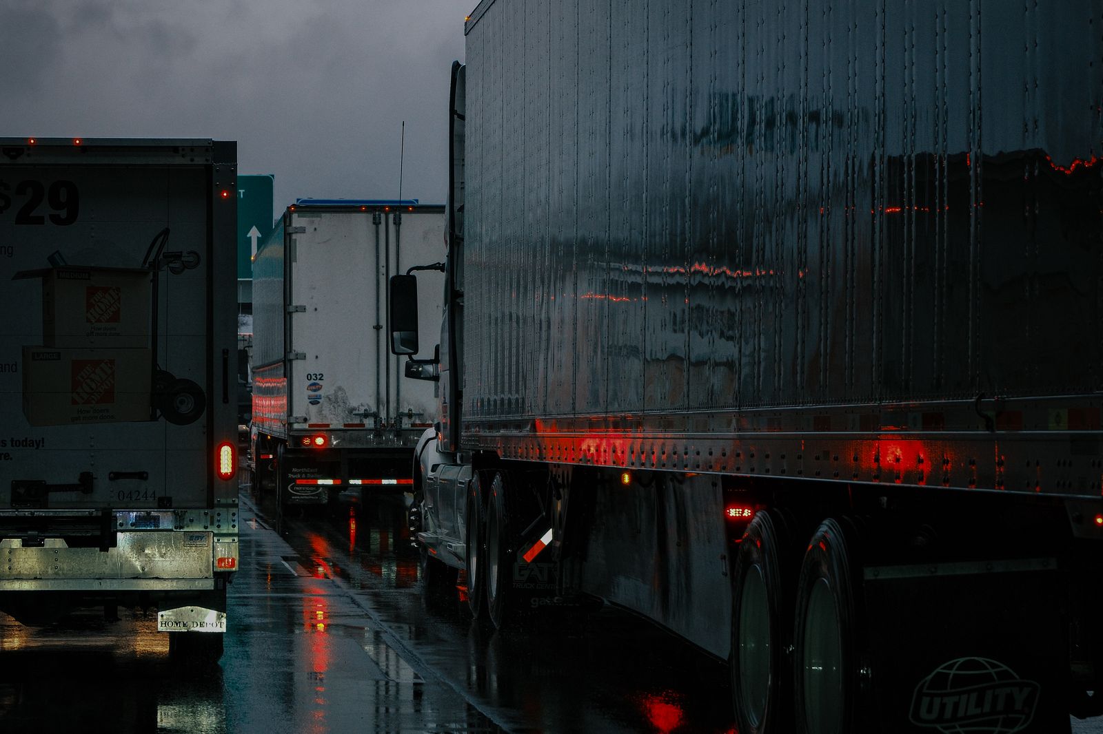 Trucks lined up on a rainy highway at night, brake lights reflecting off wet pavement