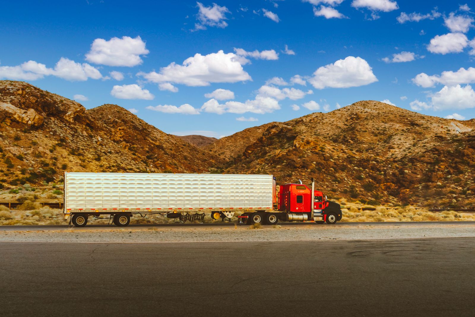 A red semi truck rolling down a desert highway with mountains on the horizon